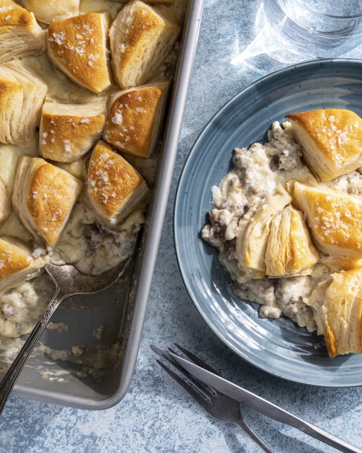 Biscuits and Gravy Casserole, with a creamy chicken with white gravy bottom and biscuits baked across the top in a baking tin, with a piece cut out on a blue plate on the side.