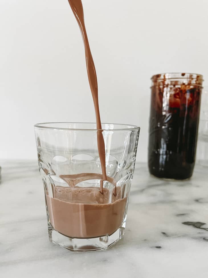 Chocolate milk being poured into a glass, with a ball jar with chocolate sauce in the background, on a marble surface.