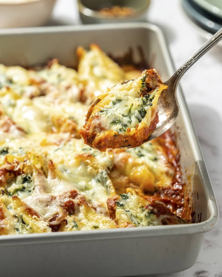 Spinach stuffed shells in a baking tin, with a piece being lifted by a silver serving spoon.