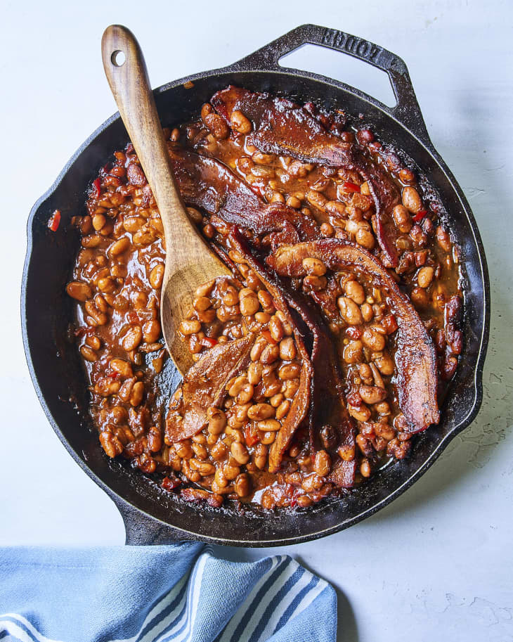 Baked beans with strips of bacon on top, in a cast iron skillet, with a wooden spoon, with a blue napkin on the side.