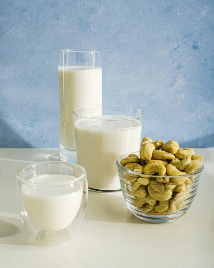 Three glasses of cashew milk (white in color) in glassware of staggered sizes, against a blue background, with bowl of cashews in a clear glass bowl next to them.