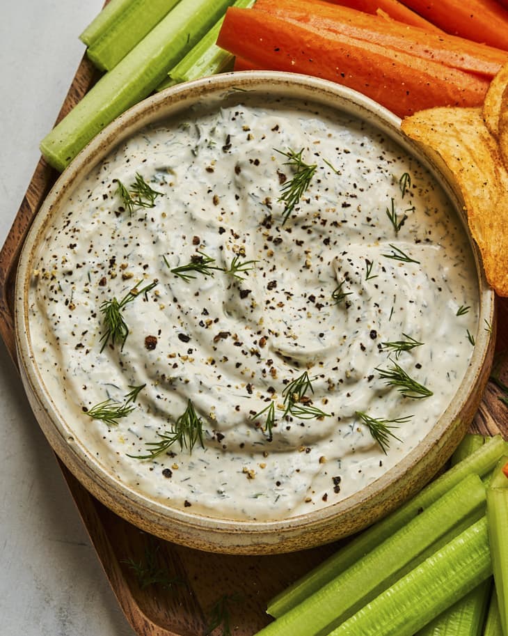a bowl of creamy dill dip, on a plate with sliced carrots, celery and potato chips