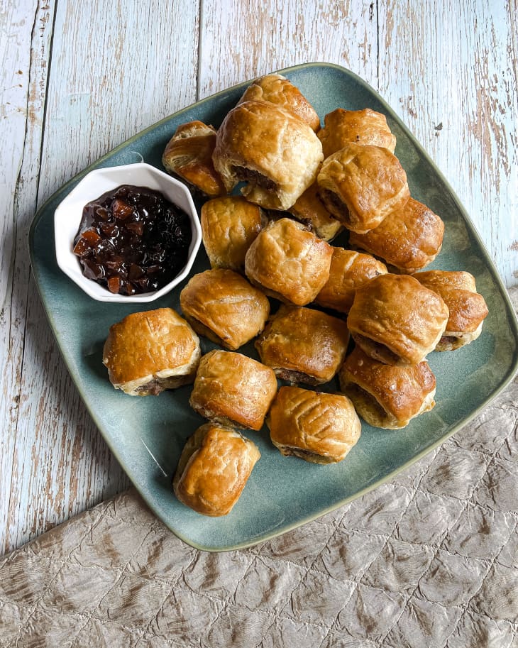 a platter of small sausage rolls on a square turquoise plate with a brownish-red dipping sauce