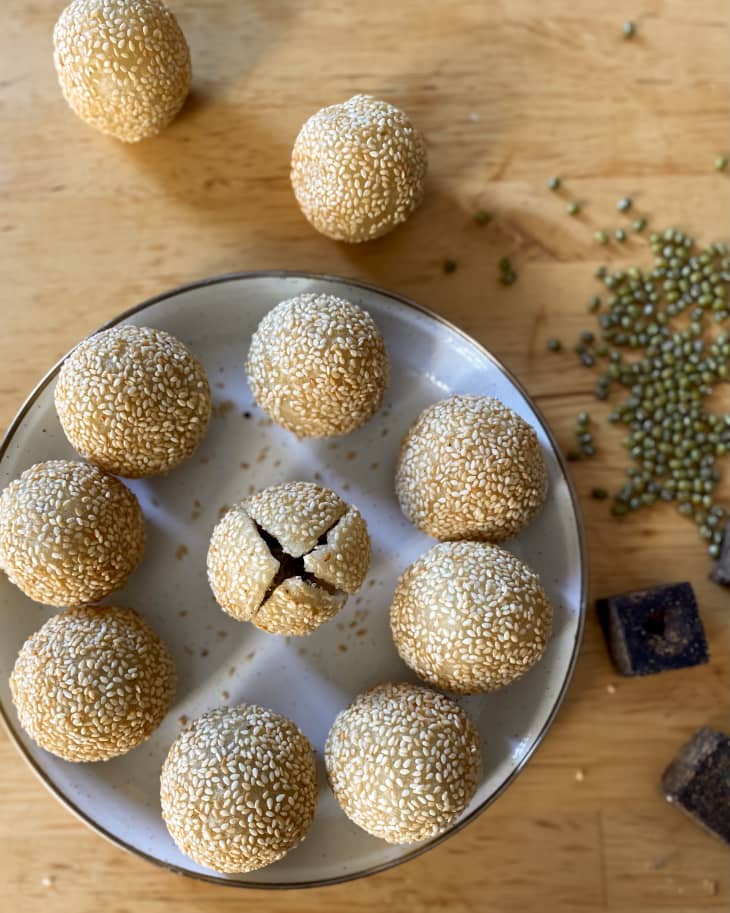 A plate with sesame balls (made with a sticky rice flour dough, filled with a sweet paste, rolled in sesame seeds, and fried until crispy on the outside)  with two more sesame balls on the table next to plate