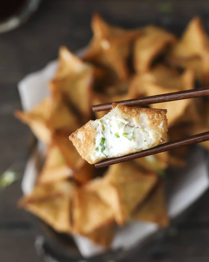 a crispy cream cheese wonton with a bite taken out so that you can see the inside, being held up by dark wood chopsticks, with the plate with the rest of the wontons in the background