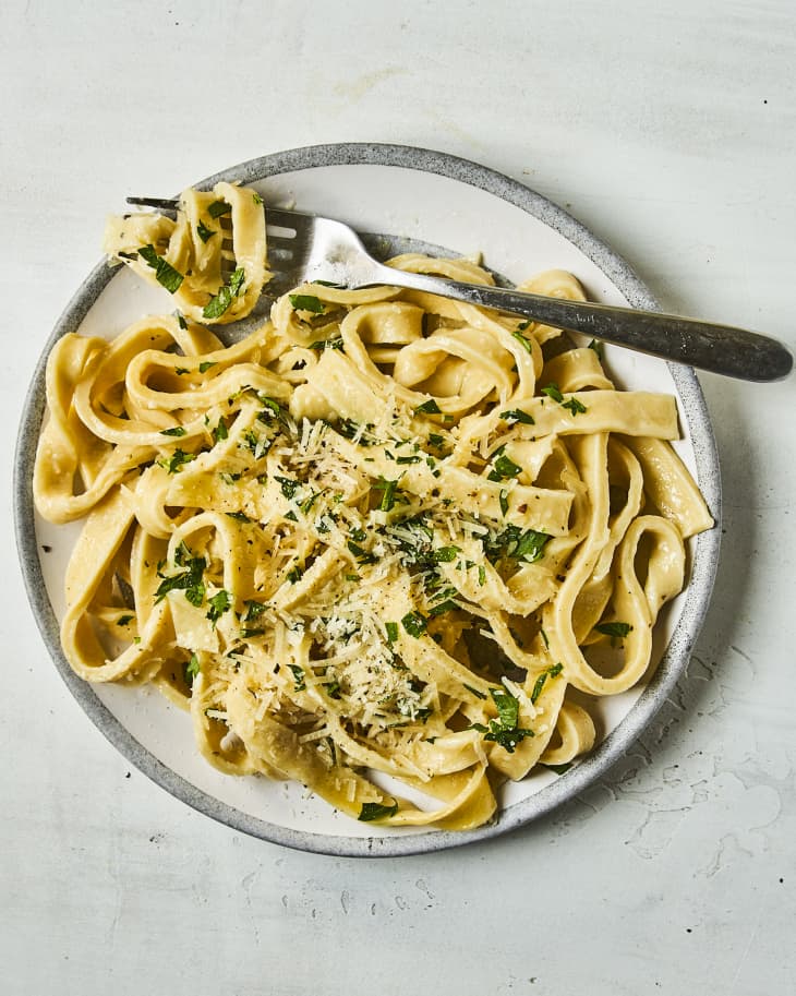 cooked egg noodles (Egg noodles are noodles made with flour and eggs. The dough is rolled thin, cut into strips, and cooked in boiling water) topped with grated parmesan cheese and parsley on a gray plate with a fork in it.