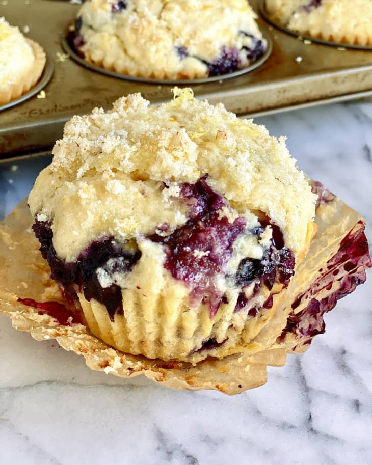 A lemon blueberry muffin with the wrapper pulled off of the sides, and the baking tin with the rest of the muffins in the background.