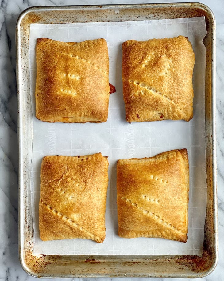 four pizza pockets (dough pockets with pizza fillings inside) on a silver baking tray with white parchment paper underneath