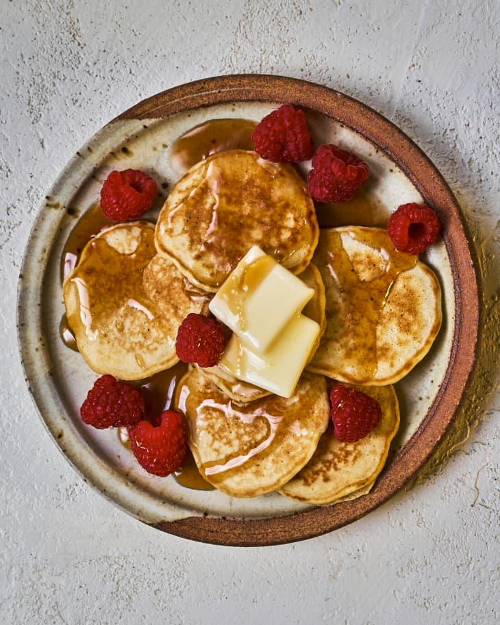 Silver dollar pancakes ona. plate with two pats of butter, raspberries and maple syrup drizzled on top.
