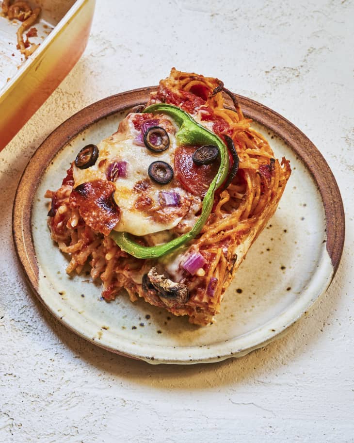 a square slice of baked spaghetti with melted cheese and pizza toppings (green peppers, onions and black olives) on top on a round ceramic plate