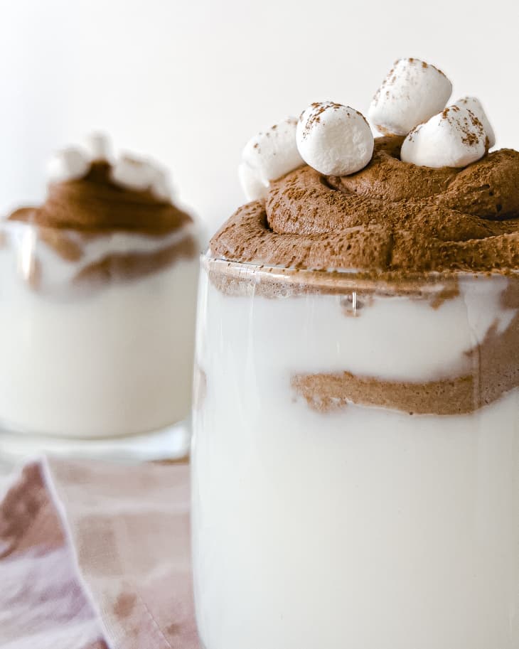 two clear glass mugs with white drinks with chocolate shavings and marshmallows on top.