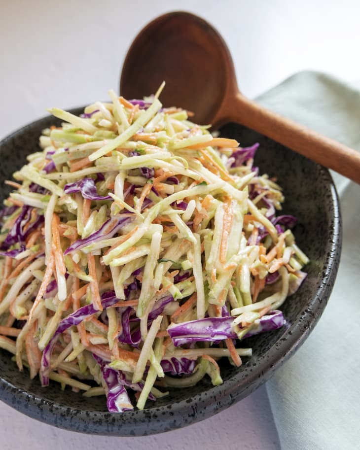 a bowl of cole slaw made with broccoli (broccoli, cabbage, carrot and a mayo based dressing, julienned and tossed together) in a black, round, bowl, with a wooden spoon on the side.