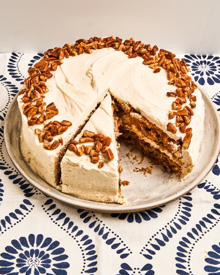 a whole, round, vegan carrot cake with white icing and nuts around the rim, with one slice removed and another cut, on a plate, on a blue and white patterned background.