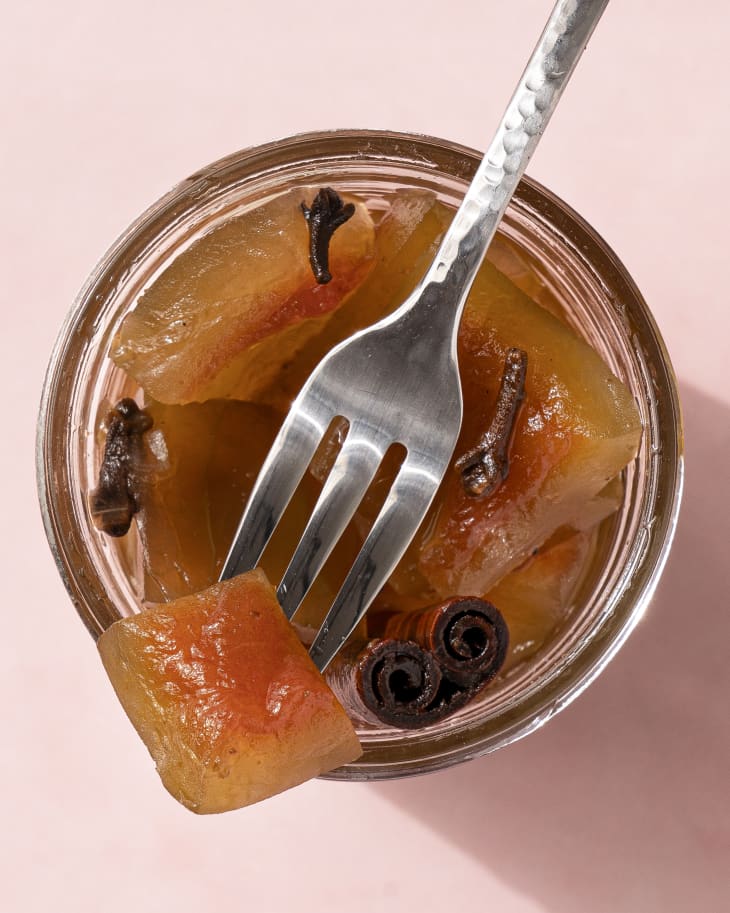 Pickled watermelon rind in a small glass jar, with a small silver fork with a piece watermelon rind balanced on the rim.
