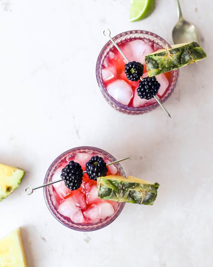 two Rum Runner's (a drink consisting of rum, banana liqueur, and grenadine) that is bright reddish pink, in ornate glasses as seen from above. There is a pineapple garnish on the rim and a glass toothpick with two blackberries balanced across the top of the glasses.