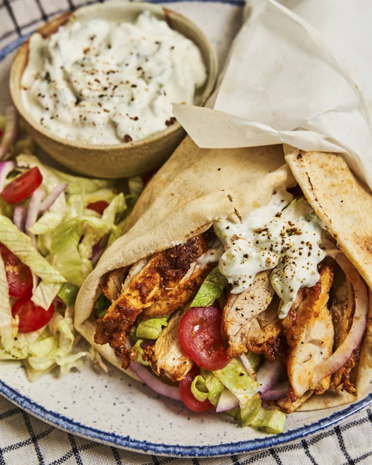 a chicken gyro on a plate, with a ramekin of tzatziki sauce and some shredded lettuce and diced tomatoes next to it.