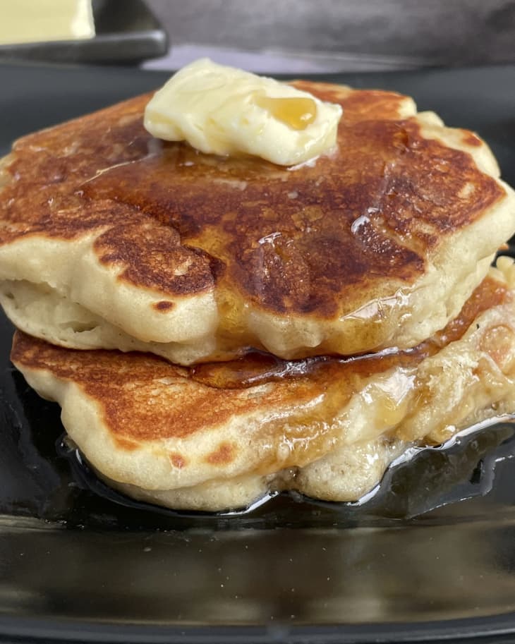 a closeup photo of two thick griddle cakes with a pat of butter on top, on a square black plate.