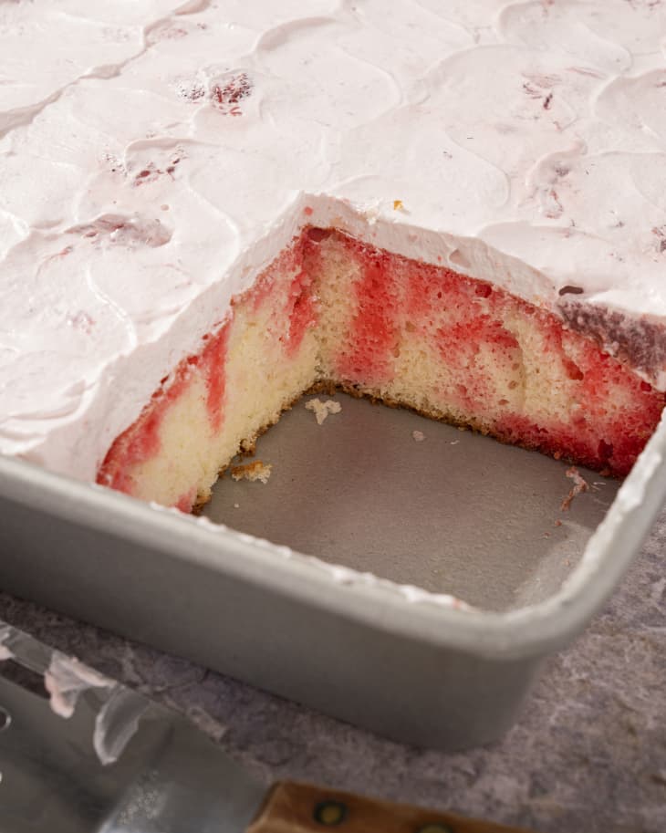 A whole sheet of vanilla cake with jello drizzles poked throughout, topped with a smooth layer of whipped cream, with a square piece from the corner of the cake cut out.