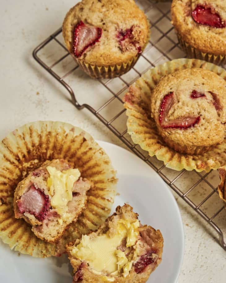 a strawberry muffin on a plate, broken in half, with more muffins on a wire cooling rack in the background.