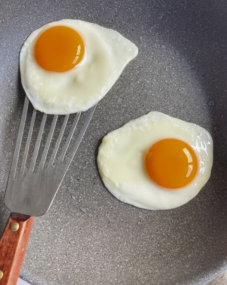Two sunny side up eggs in a gray frying pan with a metal spatula starting to lift one of the eggs.