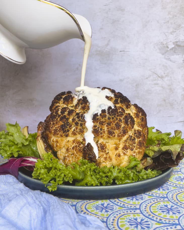 A  photo of a white sauce (Mornay sauce - a béchamel sauce with grated cheese added) being poured over a head of roasted cauliflower.