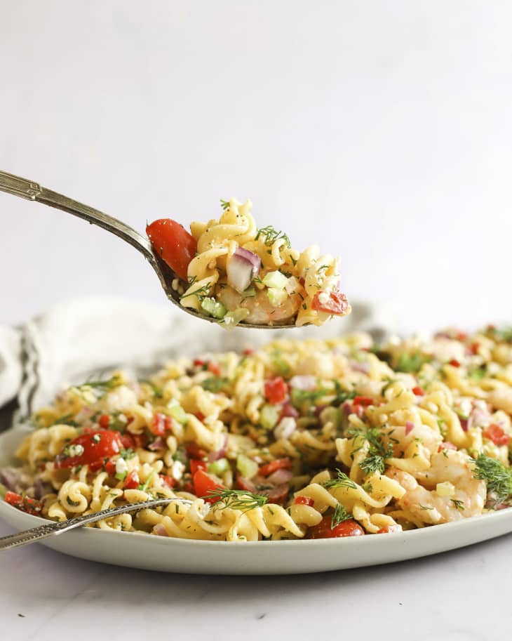 A photo of a bowl of shrimp pasta salad tossed with tomatoes and green herbs with a fork lifting a bite