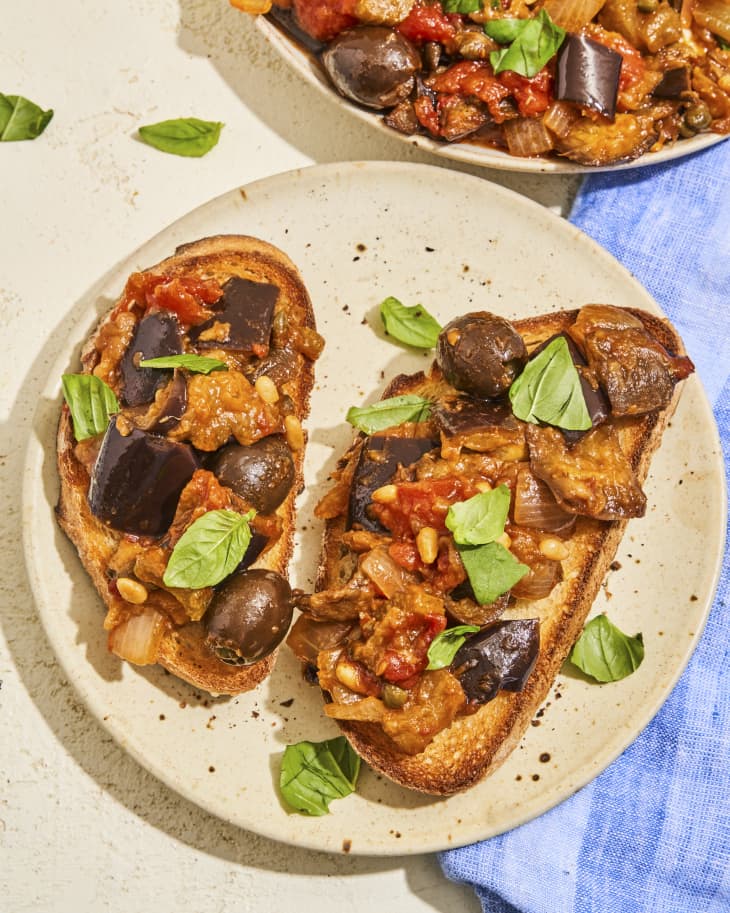 A photo of Caponata (chopped fried eggplant and other vegetables, seasoned with olive oil, tomato sauce, celery, olives, and capers) on top of two slices of toast, on a round plate, garnished with basil.