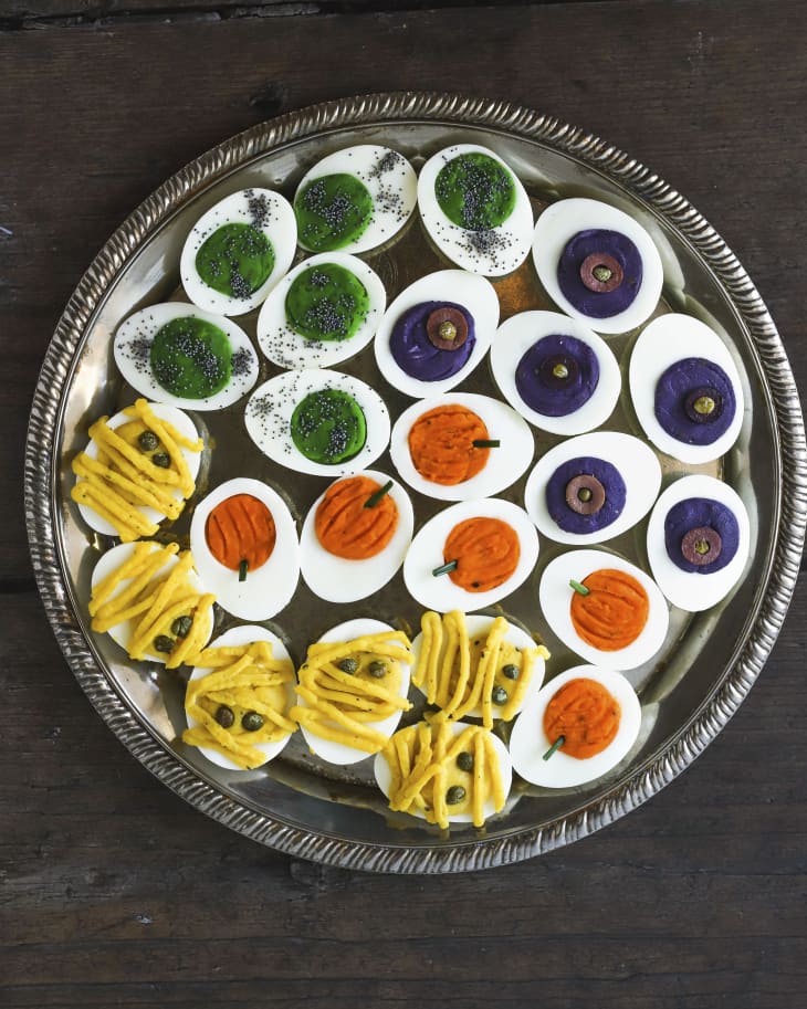 A photo of a round platter of deviled eggs, with the yolk filling made into different colors with various halloween figures such as pumpkin and mummies.