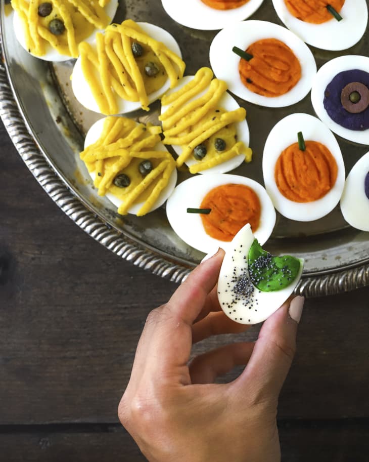 A photo of a round platter of deviled eggs, with the yolk filling made into different colors with various halloween symbols such as pumpkin and mummies, with a hand holding a deviled egg with a green yolk filling.