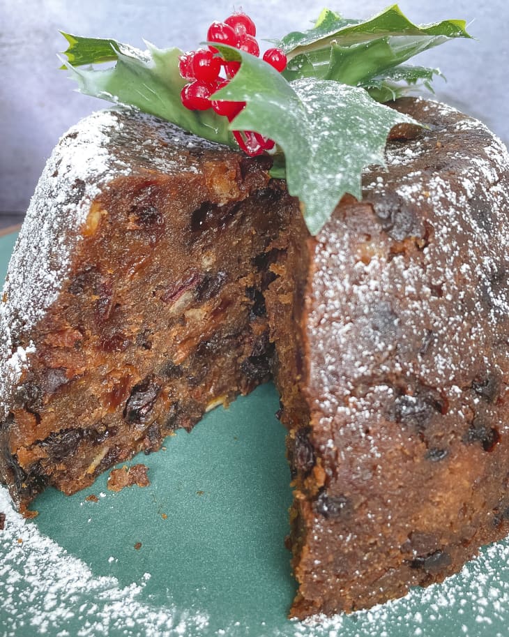 A photo of a whole Christmas Pudding/Figgy pudding (a traditional Christmas round cake made with dried fruit) topped with a holly sprig and powder sugar with a slice cut out.