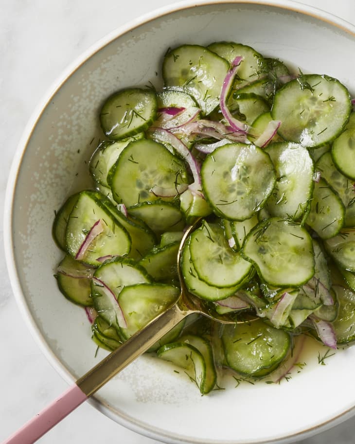 A bowl of cucumber onion salad on a marble surface with a pink-handled spoon reaching in for a scoop.