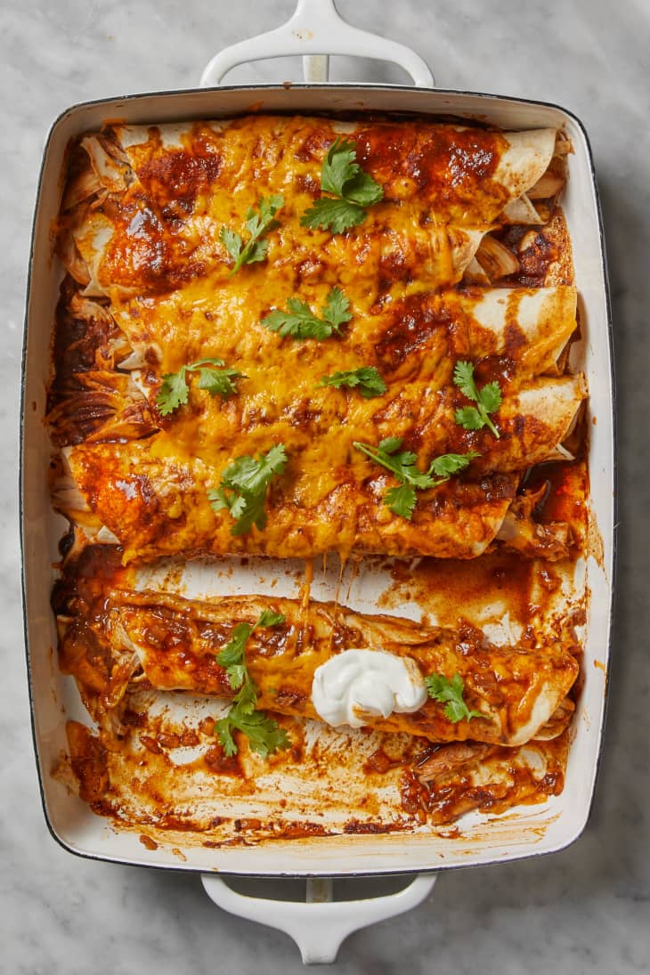 An overhead view of a white baking dish with turkey enchiladas covered in melted cheese, sauce, and cilantro on a white marble countertop, with one enchilada removed