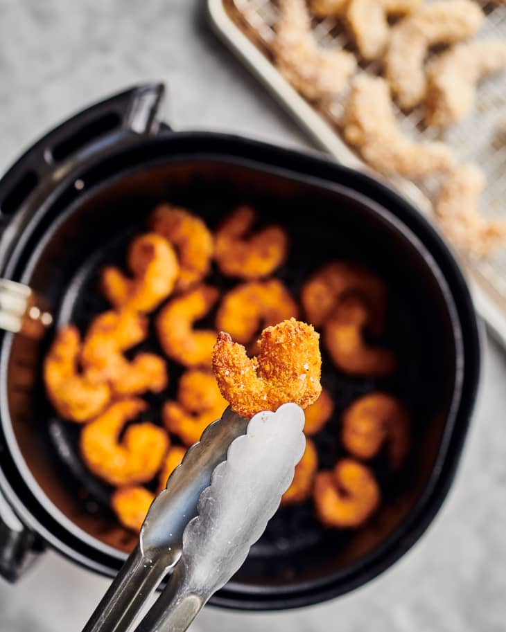 tongs are holding one piece of fried shrimp out of an air fryer with fried shrimp in it
