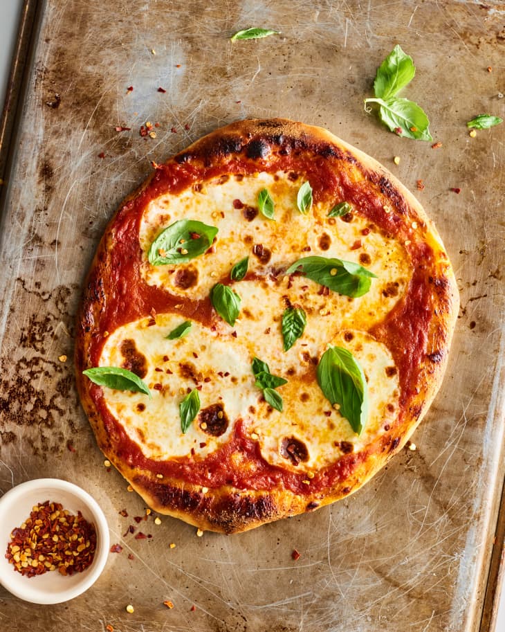 Margherita pizza with fresh basil and mozzarella on a baking sheet, accompanied by a small bowl of red pepper flakes.