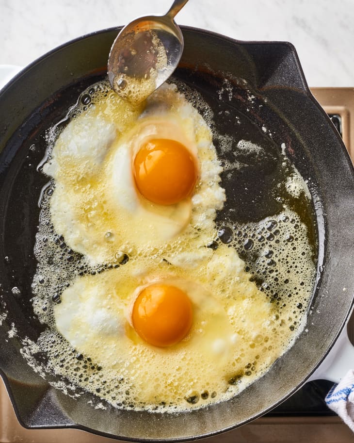 Someone scoops butter from a skillet and puts it on fried eggs  while cooking