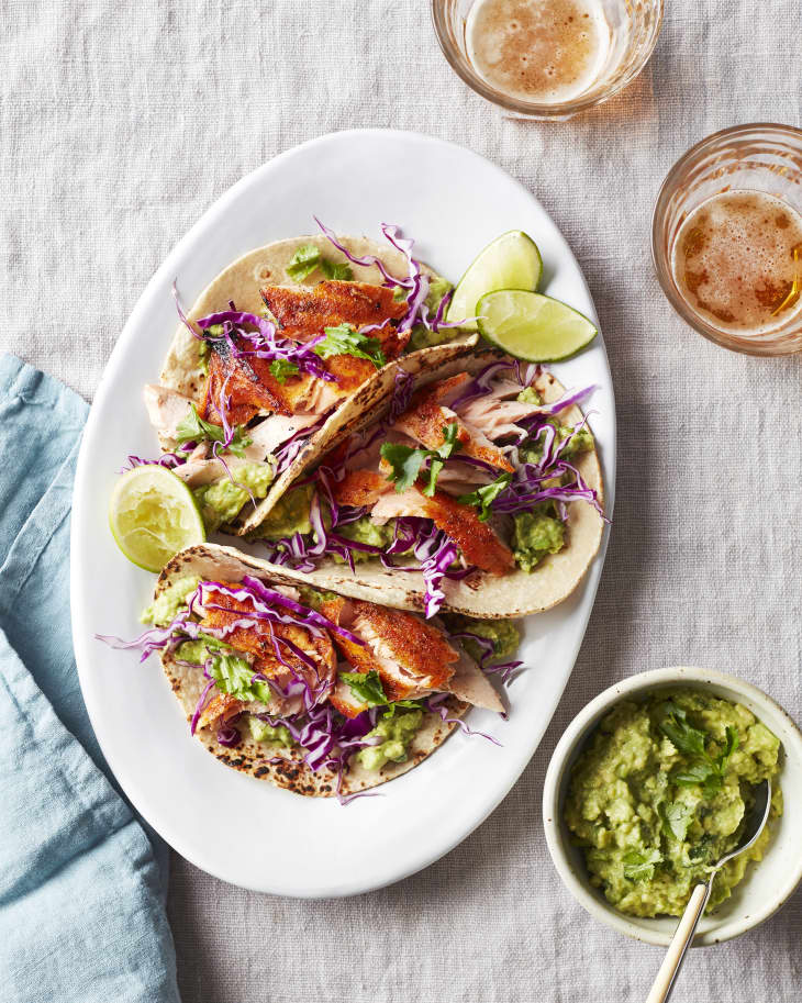 A platter of chili-lime salmon tacos sits on a table next to a two drinks and guacamole.