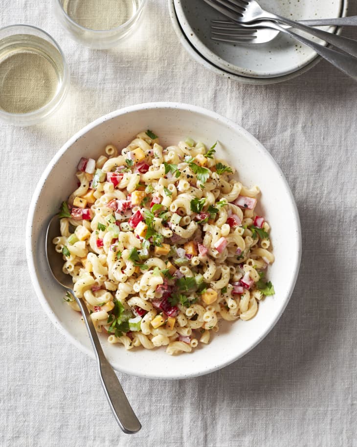 A large bowl of macaroni salad sits on kitchen table next to glasses of wine, bowls, and forks.