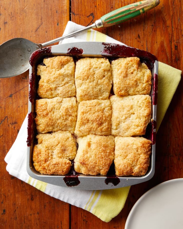 The baked berry cobbler sits on top of a linen. A spoon lays off to the side of the pan.