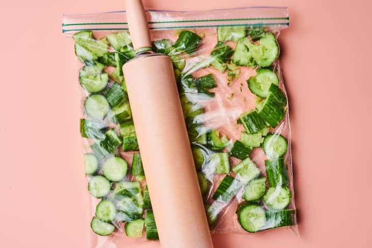 A wooden rolling pin smashes cut up cucumbers in a plastic bag.
