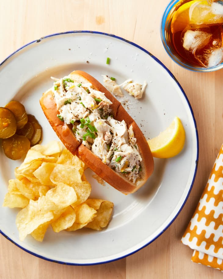 Crab salad garnished with chives in brioche hotdog bun. Potato chips and sliced pickled on the side. Iced tea with lemon and graphic tea towel in the background.