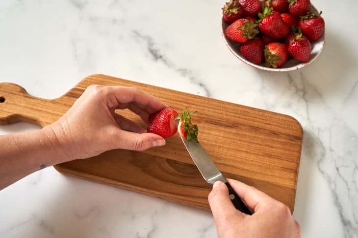 someone holding strawberry in left hand with knife in right hand removing top from strawberry