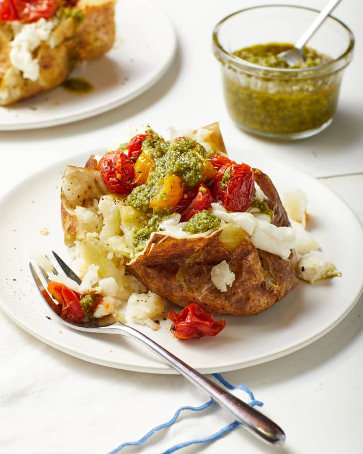 Summer baked potato on a white plate with fork on the side. Baked potato is topped with ricotta, blistered yellow and red cherry tomatoes, and green pesto. Glass ramekin filled with pesto in the background