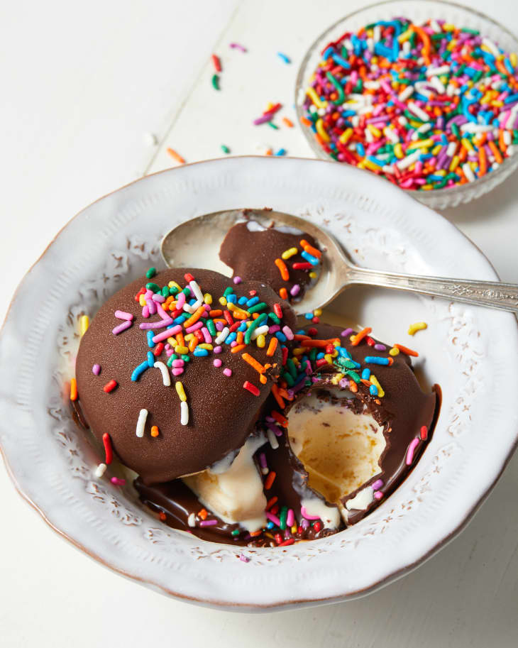 Vintage white bowl with two scoops of vanilla ice cream topped with chocolate magic shell and multi-colored sprinkles.  Someone has taken a scoop of ice cream and placed spoon on the side