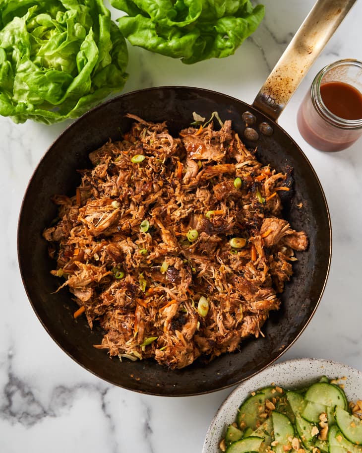 Shredded pork topped with sliced scallion in black skillet. In background Boston lettuce, pickles and jar of barbecue sauce.