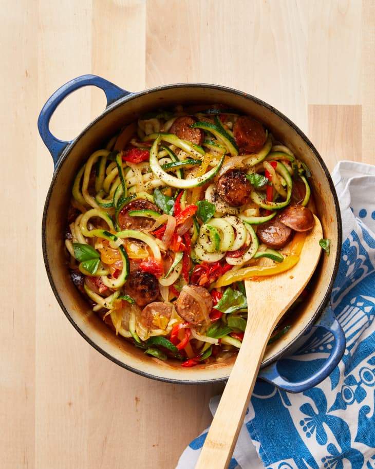 Blue Dutch oven filled with zucchini noodles, sausage and peppers; wooden spoon rest on inside of pot