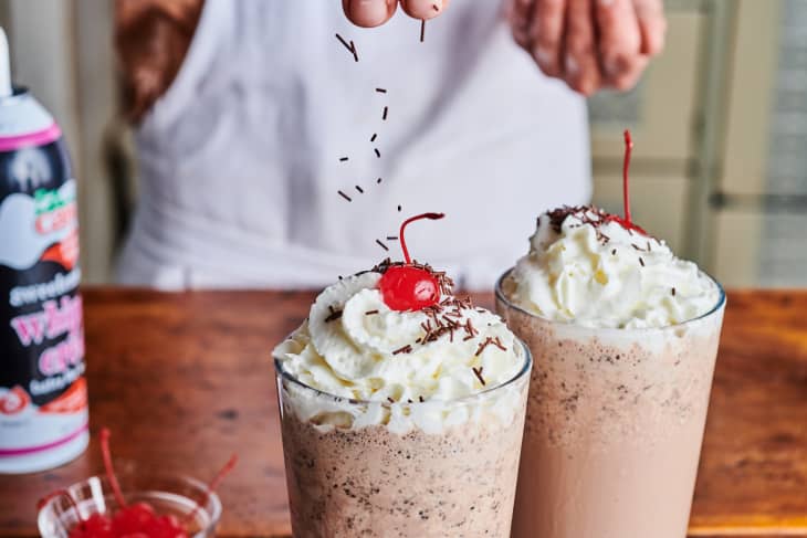 Someone adds sprinkles over top of the cherry and whip cream topped chocolate milkshake.