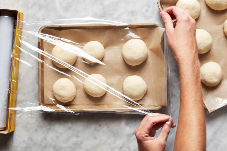 Seran wrap is pulled over a payment lined baking sheet with six dough balls.