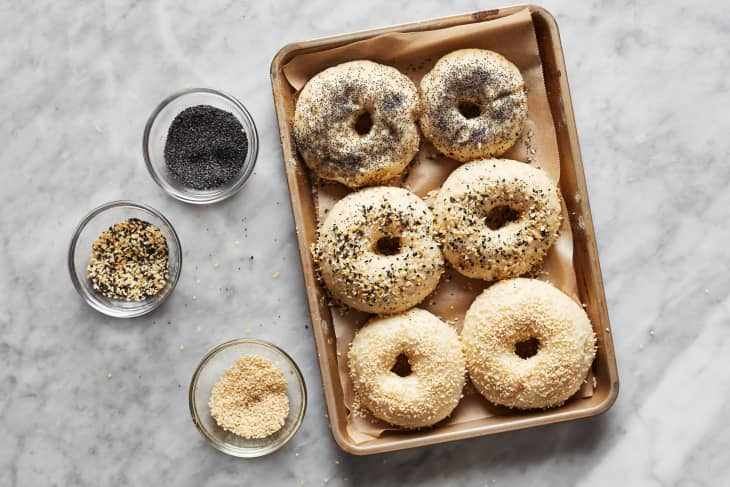 A sheet pan of six unbaked bagels sit on a counter. A small ramekin of white sesame seeds, a small ramekin of black sesame seeds, and a small ramekin of everything bagel seasoning sit off to the side.