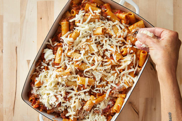 Rigatoni and beef being topped with shredded cheese in baking dish.