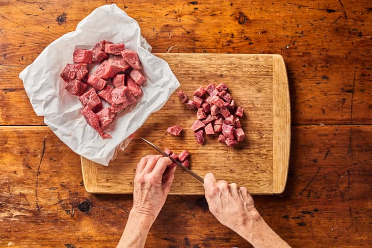 someone is chopping raw beef on a cutting board with larger chunks separated near it in parchment paper